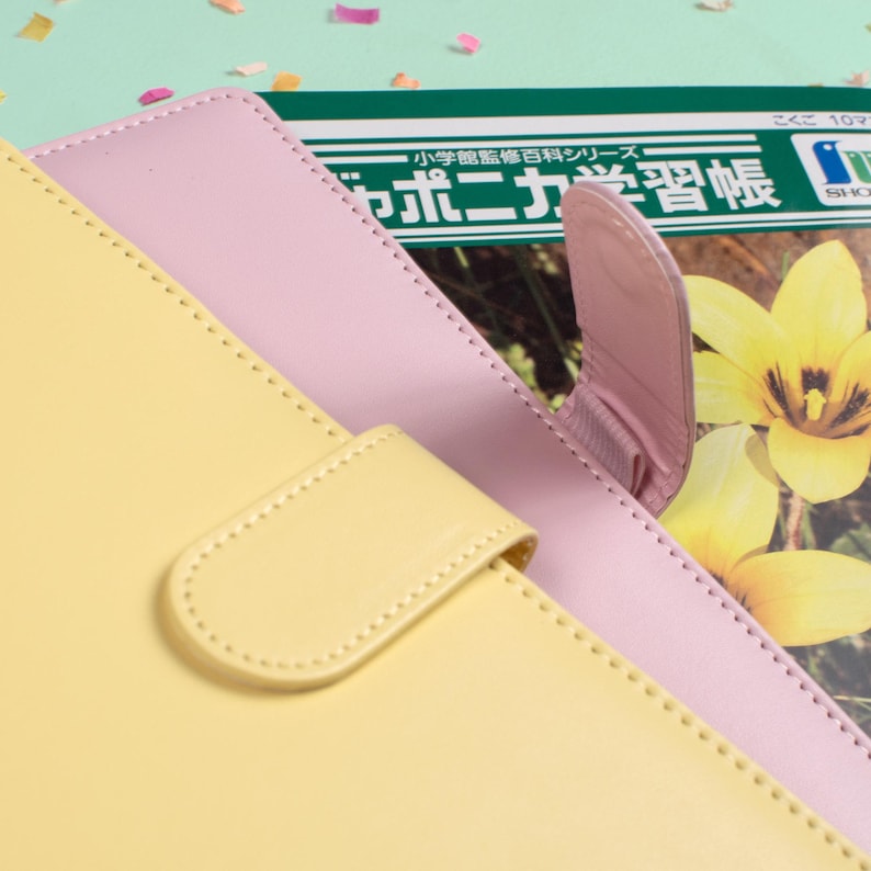 Peut inclure: Gros plan sur un carnet en cuir jaune pastel et rose avec un fermoir assorti. Le carnet est partiellement ouvert, r&eacute;v&eacute;lant un aper&ccedil;u d'un manuel japonais avec des fleurs jaunes en arri&egrave;re-plan. La couverture a une bordure cousue.