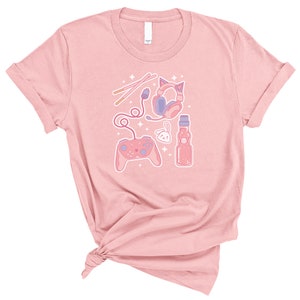 K&ouml;nnte beinhalten: Pinkes T-Shirt mit einem Grafikdesign aus einem rosa und violetten Videospiel-Controller, Kopfh&ouml;rern, einer Flasche Ramune-Soda und einem rosa Hasen. Das Design ist von wei&szlig;en Sternen umgeben.