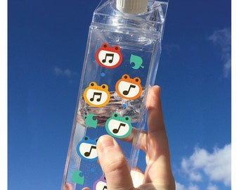 May include: A clear plastic milk carton-shaped water bottle with colorful frog stickers featuring musical notes. The bottle is held in a hand against a blue sky background.