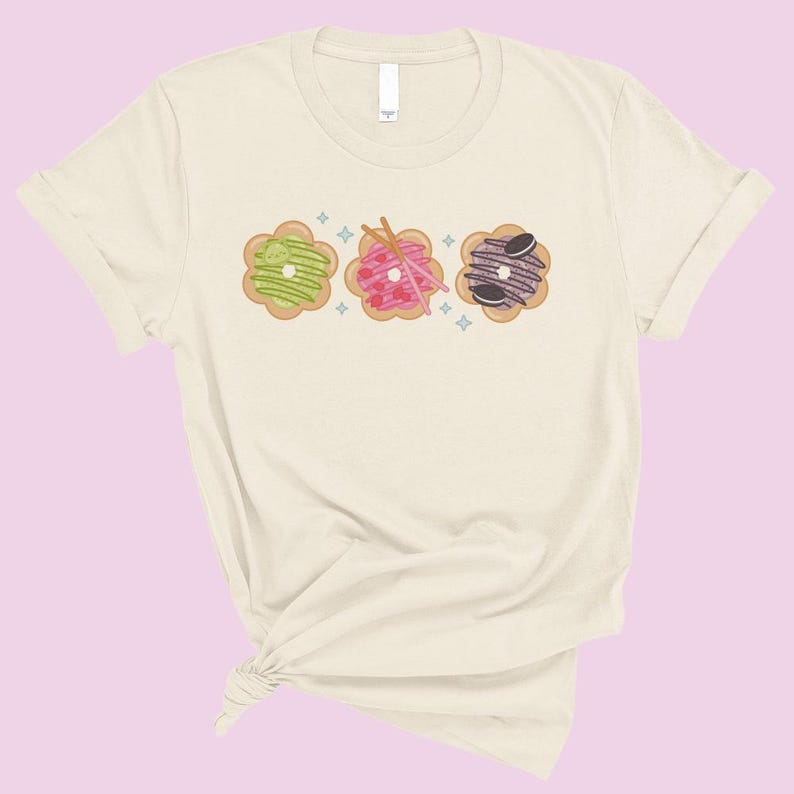 May include: A cream-colored t-shirt with a graphic of three colorful donuts with different toppings. The donuts are arranged in a row and have a whimsical, cartoon-like style.