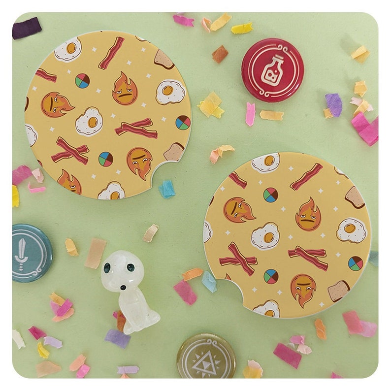May include: Two yellow car coasters with a breakfast food pattern, including bacon, eggs, and toast, and various emoji faces. Also, several decorative pins and a small figurine are scattered on a light green surface.