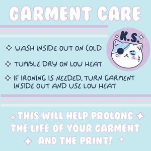 May include: A light blue and pink graphic with the text "GARMENT CARE" in white. The graphic includes instructions for washing, drying, and ironing garments. A cartoon cat with a pirate eye patch and the text "K.S." is in the upper right corner. The text reads: "WASH INSIDE OUT ON COLD", "TUMBLE DRY ON LOW HEAT", "IF IRONING IS NEEDED, TURN GARMENT INSIDE OUT AND USE LOW HEAT", "THIS WILL HELP PROLONG THE LIFE OF YOUR GARMENT AND THE PRINT!"