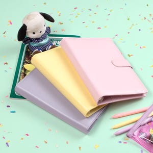 Peut inclure: Trois carnets de couleurs pastel : lavande, jaune et rose. Un chien en peluche est pos&eacute; sur un livre vert. Plusieurs crayons de couleur et une petite trousse sont visibles sur une surface vert clair, avec des confettis &eacute;parpill&eacute;s.