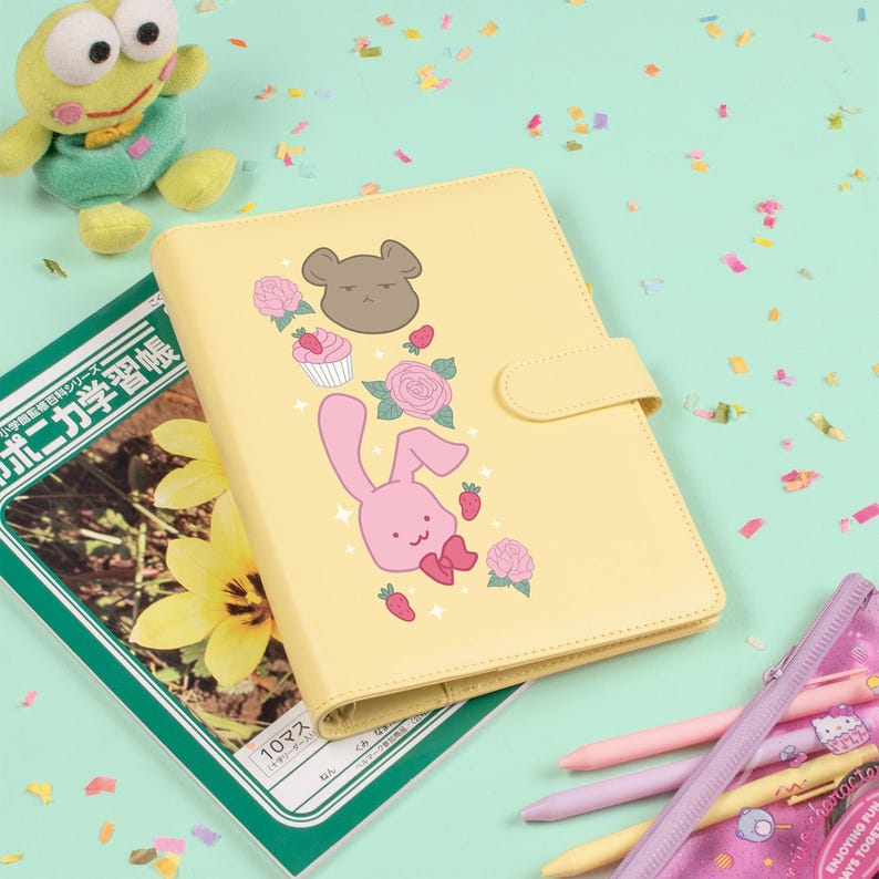 Peut inclure: Un agenda jaune clair avec un motif de lapin rose, d'ours, de cupcake et de roses. L'agenda a une fermeture &agrave; bouton-pression et se trouve &agrave; c&ocirc;t&eacute; d'un livre vert et d'un jouet grenouille en peluche. Des stylos et une trousse sont &eacute;galement dans l'image.
