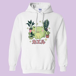 May include: White hoodie with a green frog sitting among potted plants. The frog is smiling and has a pink tongue sticking out. The text "カエル" is written in pink below the frog.