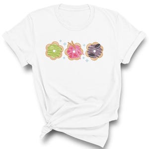 May include: White t-shirt with a graphic of three colorful donuts with different toppings.