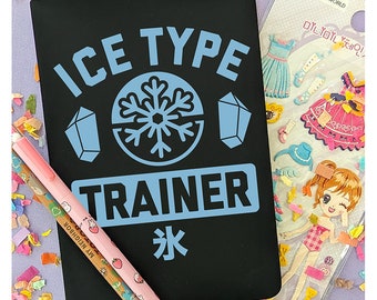 May include: Black notebook with the text "ICE TYPE TRAINER" in white and blue lettering. A snowflake graphic is in the center of the notebook. The notebook is surrounded by colorful confetti and stickers.