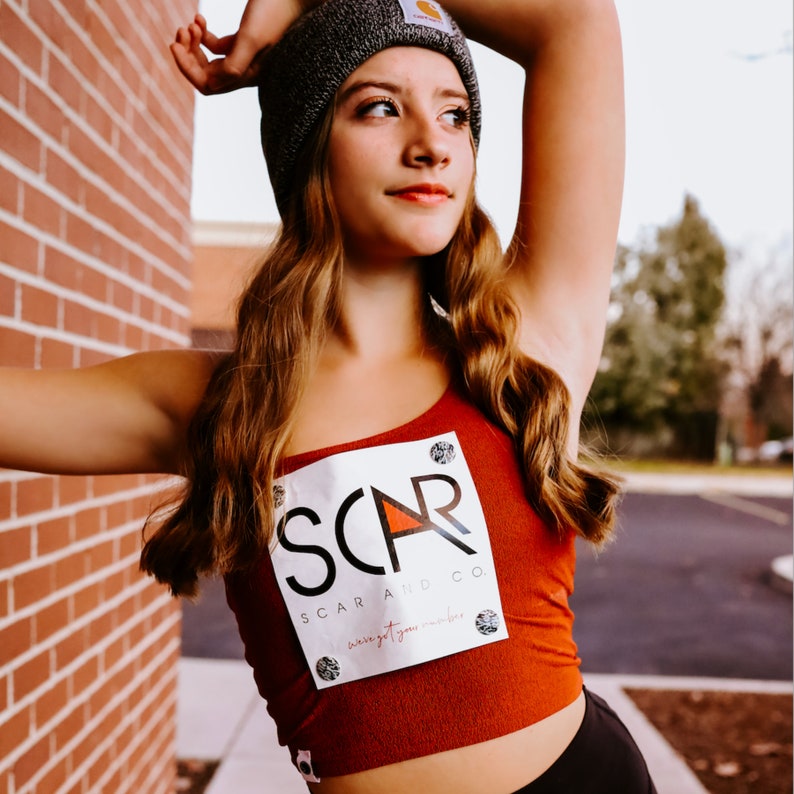 Op de afbeelding: Een jong persoon draagt een rood tanktop met een witte vierkante patch waarop staat "SCAR AND CO. swing at your number". De persoon draagt een zwarte beanie met een wit logo en heeft lang bruin haar.