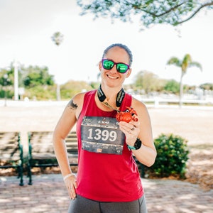 May include: A woman wearing a red tank top and grey leggings is holding a medal with the number 1399 on it. She is smiling and wearing sunglasses.