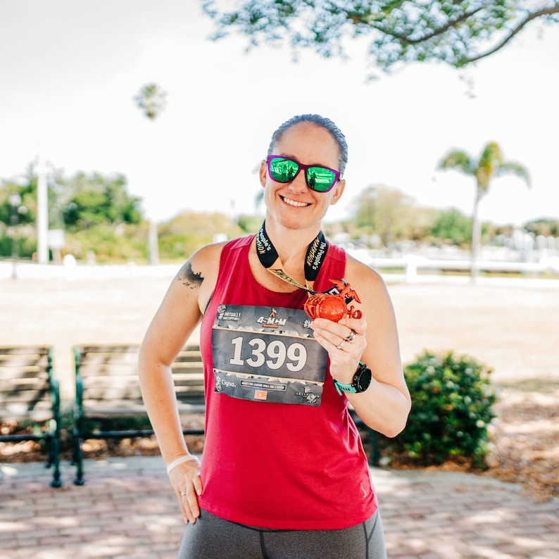 May include: A woman wearing a red tank top and grey leggings is holding a medal with the number 1399 on it. She is smiling and wearing sunglasses.