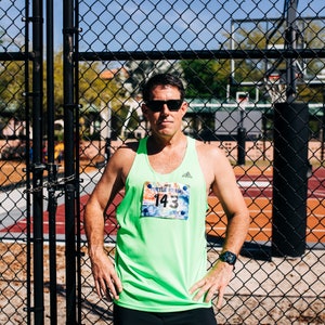 Op de afbeelding: Een man in een groen tanktop met een wedstrijdnummer waarop "2015 Turtle Crawl 143" staat, staat voor een hek van gaas.