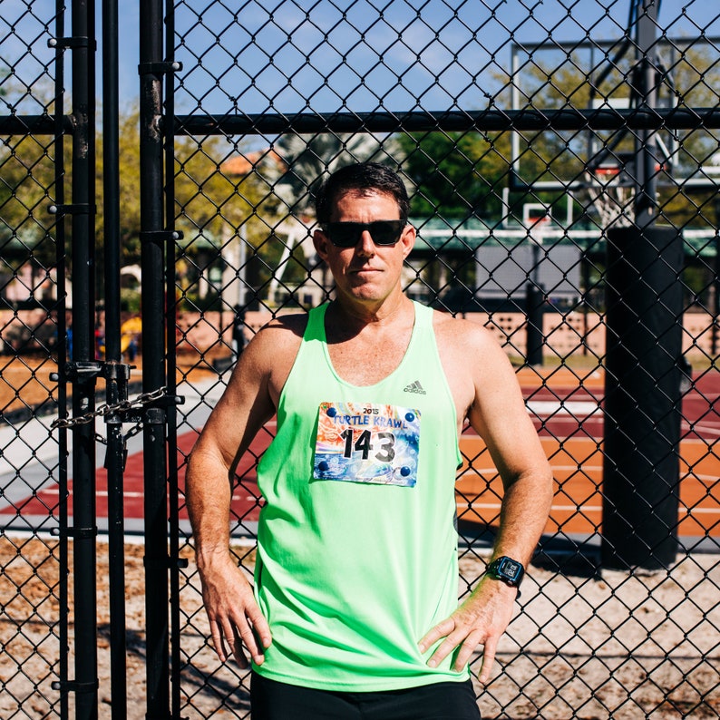 Op de afbeelding: Een man in een groen tanktop met een wedstrijdnummer waarop "2015 Turtle Crawl 143" staat, staat voor een hek van gaas.