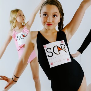 Op de afbeelding: Twee meisjes dragen maillotjes met het logo "SCAR AND CO" erop gedrukt. Het maillotje links is roze met ruches en het maillotje rechts is zwart.
