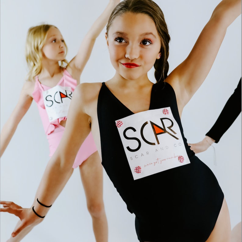 Op de afbeelding: Twee meisjes dragen maillotjes met het logo "SCAR AND CO" erop gedrukt. Het maillotje links is roze met ruches en het maillotje rechts is zwart.
