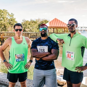May include: Three men wearing green and blue shirts with race numbers, standing outdoors near a body of water. The men are wearing sunglasses and shorts. The man in the middle has a black and blue shirt with the number 748. The man on the left has a green tank top with the number 143. The man on the right has a green long-sleeved shirt with the number 61.