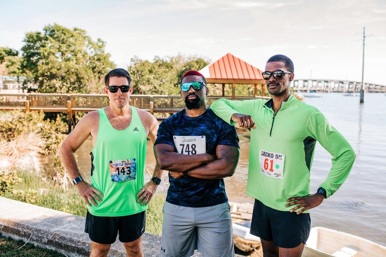 May include: Three men wearing green and blue shirts with race numbers, standing outdoors near a body of water. The men are wearing sunglasses and shorts. The man in the middle has a black and blue shirt with the number 748. The man on the left has a green tank top with the number 143. The man on the right has a green long-sleeved shirt with the number 61.