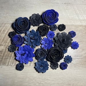 May include: A collection of blue and black paper flowers arranged in a circle on a woodgrain background. The flowers are all different sizes and shapes, and some have a more intricate design than others.
