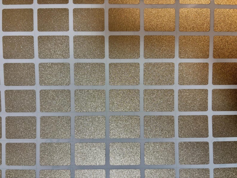 May include: Close-up of a sheet of gold glitter stickers. The stickers are rectangular with rounded corners and are arranged in a grid pattern. The background is white.