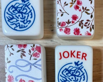 May include: Four white rectangular mahjong tiles. Two tiles feature the red text JOKER above a blue circular dragon emblem. The other two tiles display a floral pattern with pink roses, green leaves, and thin brown stems on a white background.
