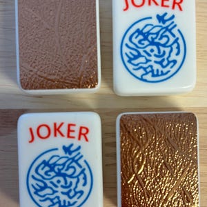 Może przedstawiać: Cztery kafelki do gry w Mahjonga. Dwa białe kafelki ze słowem "JOKER" w kolorze czerwonym i niebieskim okrągłym wzorem. Dwa brązowe kafelki z teksturowanym wzorem. Kafelki są prostokątne z zaokrąglonymi rogami.