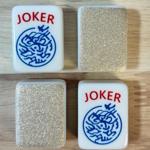 May include: Four white Mahjong tiles. Two tiles feature the word "JOKER" in red and a blue design. The other two tiles have a glittery gold surface. The tiles are rectangular with rounded corners.