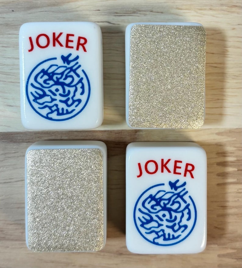 May include: Four white Mahjong tiles. Two tiles feature the word "JOKER" in red and a blue design. The other two tiles have a glittery gold surface. The tiles are rectangular with rounded corners.