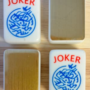 Op de afbeelding: Vier witte Mahjong-tegels. Twee tegels tonen het woord "JOKER" in het rood boven een blauw cirkelvormig ontwerp. De andere twee tegels hebben een goudkleurig oppervlak. De tegels zijn rechthoekig met afgeronde hoeken.