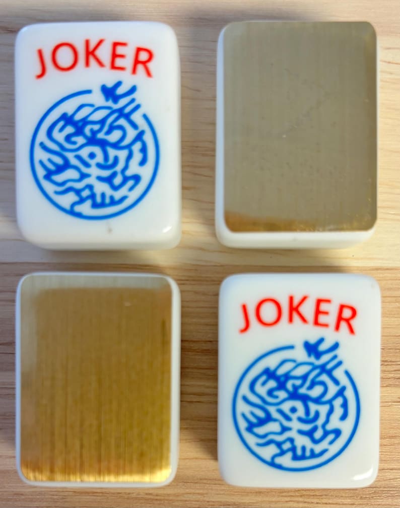 Op de afbeelding: Vier witte Mahjong-tegels. Twee tegels tonen het woord "JOKER" in het rood boven een blauw cirkelvormig ontwerp. De andere twee tegels hebben een goudkleurig oppervlak. De tegels zijn rechthoekig met afgeronde hoeken.