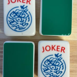 May include: Four white and green Mahjong tiles. Two tiles read "JOKER" in red, with a blue circular design. The other two tiles are solid green. The tiles are rectangular with rounded corners.