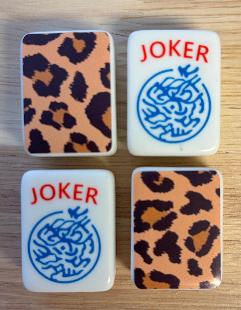 May include: Four rectangular Mahjong tiles. Two tiles feature a leopard print pattern in brown and orange. The other two tiles are white with the word "JOKER" in red and a blue circular design.