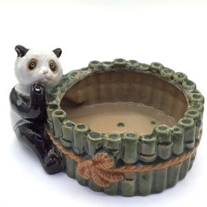 Könnte beinhalten: Ein Keramik-Pflanztopf in Form eines Bambus-Zaunts mit einer Panda-Figur, die auf der Seite sitzt. Der Topf ist grün und braun mit einem Seildetail am oberen Rand.