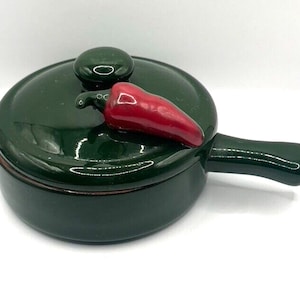 May include: A green ceramic casserole dish with a red chili pepper on the lid. The dish has a handle and a round shape.