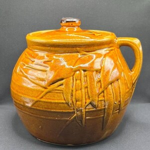 Può includere: Un vaso di ceramica smaltata con coperchio, manico e base marrone scuro. Il vaso è di un caldo colore marrone terroso con dettagli di smalto strutturato e gocciolante. Il coperchio ha un piccolo pomello marrone scuro.