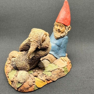 May include: A ceramic figurine of a gnome wearing a red hat and blue shirt, sitting next to a brown rabbit figurine. The figurines are on a base with a brown, orange, and yellow leaf design.