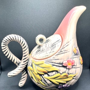 May include: A decorative, ceramic teapot with a unique, curved design. The teapot is primarily pink with a textured pattern and features a curved handle with black and white stripes. It is adorned with yellow leaves and colorful flowers.