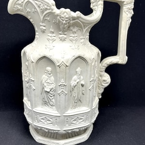 May include: White ceramic pitcher with an ornate design featuring religious figures in relief. The pitcher has a wide, flared spout and a handle with a sculpted face.