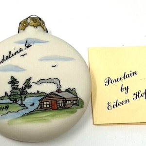 May include: A white porcelain Christmas ornament with a hand-painted scene of a cabin by a lake. The ornament is signed "Madeline Is." and "Hepp". A small tag reads "Porcelain by Eileen Hepp".