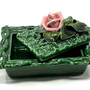 May include: A green ceramic trinket box with a lid featuring a pink rose with green leaves. The box has a textured, leafy design and a raised, rope-like border.