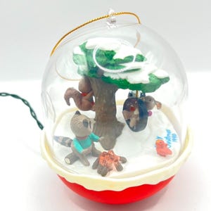May include: A clear, spherical Christmas ornament with a red base, featuring a winter scene with woodland creatures. Inside, a tree with snow, a raccoon, squirrel, and bird are visible. The ornament has a gold string for hanging and a green light string.