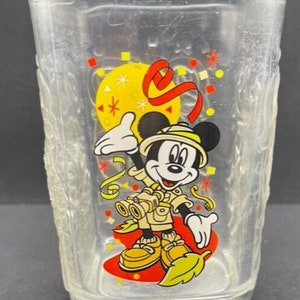 Puede incluir: Un vaso de vidrio transparente con un diseño de Mickey Mouse. Mickey lleva un sombrero de safari y sostiene unos binoculares. El vaso tiene un diseño de confeti rojo, amarillo y blanco y el año 2000 impreso en la parte inferior.