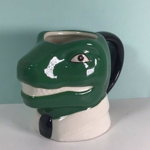 May include: A green ceramic mug shaped like a dinosaur wearing a white collar and black tie. The mug has a black handle and a single brown eye.