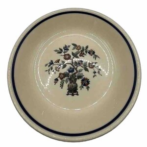 May include: A cream-colored ceramic bowl with a dark blue rim and a floral design. The design features a vase with a tree-like arrangement of flowers in shades of brown, blue, and green. The bowl is round and has a smooth surface.