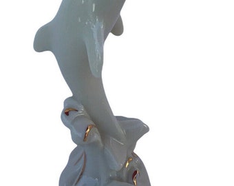 May include: White porcelain dolphin figurine with gold accents. The dolphin is leaping out of the water with its mouth open. The base of the figurine is decorated with gold swirls.