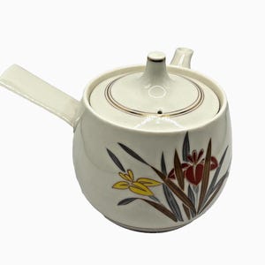 May include: A white ceramic teapot with a side handle and a matching lid. The body features a painted floral design with yellow and dark red flowers, green leaves, and grey accents. The lid has a small knob and a thin gold-colored circular rim.