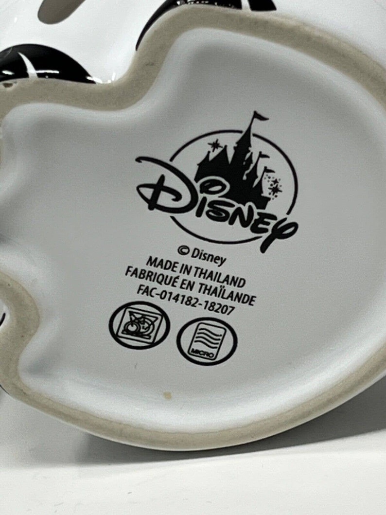 May include: White ceramic mug with a black and white Mickey Mouse design. The bottom of the mug features the Disney logo, text that reads "MADE IN THAILAND", and other symbols.