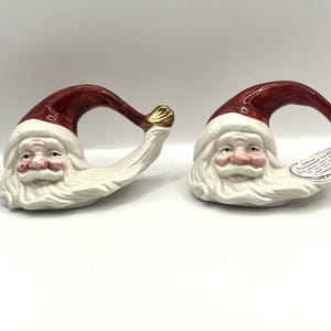 Puede incluir: Dos servilleteros de cerámica con la figura de Papá Noel. Cada servilletero presenta una cara de Papá Noel con barba blanca y gorro rojo. El gorro se curva en forma de anillo con un adorno dorado. Las caras tienen mejillas rosas.