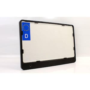 Könnte beinhalten: Ein schwarzer Kennzeichenhalter mit einem weißen Kennzeichen. Das Kennzeichen hat einen blauen Aufkleber mit gelben Sternen und dem Buchstaben "D". Der Rahmen ist rechteckig und hat eine glänzende Oberfläche.