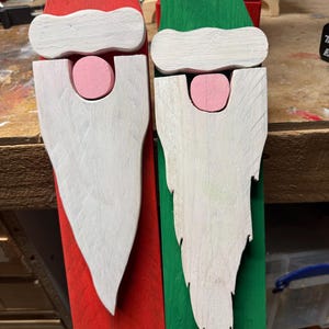 May include: Two wooden Santa Claus decorations. One is red and white, the other is green and white. Each has a white beard, a pink nose, and a white hat trim. The beards are cut in a stylized shape.