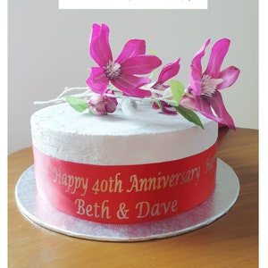 May include: A white cake with a red ribbon that says "Happy 40th Anniversary Beth & Dave". The cake is decorated with pink flowers.