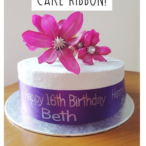 May include: A white cake with a purple ribbon that says "Happy 16th Birthday Beth". The cake is decorated with pink flowers.